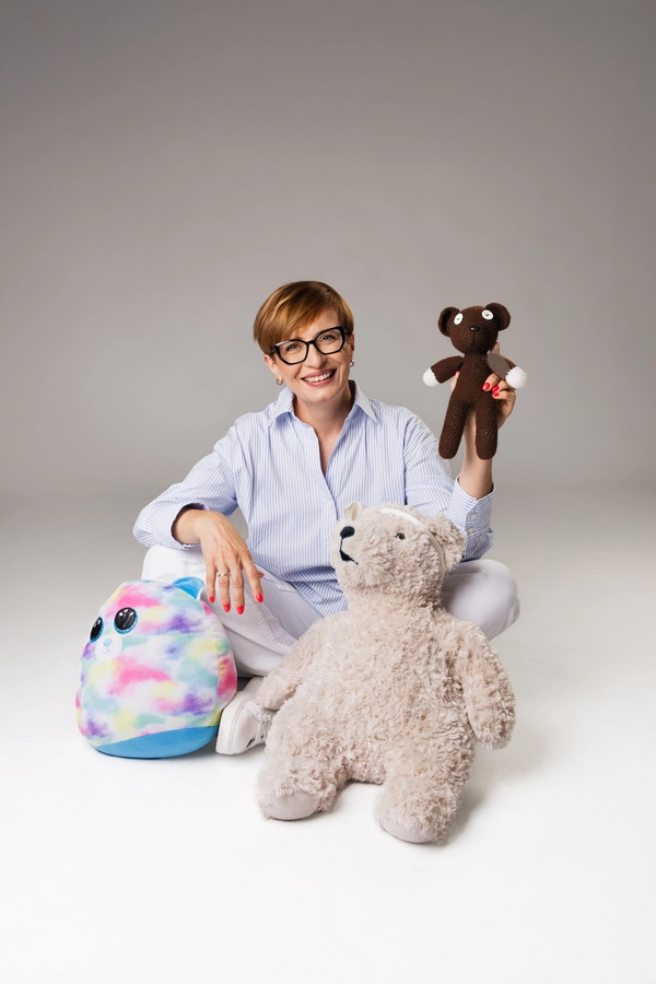 Portret Lidii Rodowicz, która siedzi na białym tle, ubrana w jasną koszulę i spodnie. Kobieta uśmiecha się, trzymając w ręku małego, brązowego pluszowego misia. Obok niej leżą duży jasny pluszowy miś i tęczowa poduszka Squishmallow.