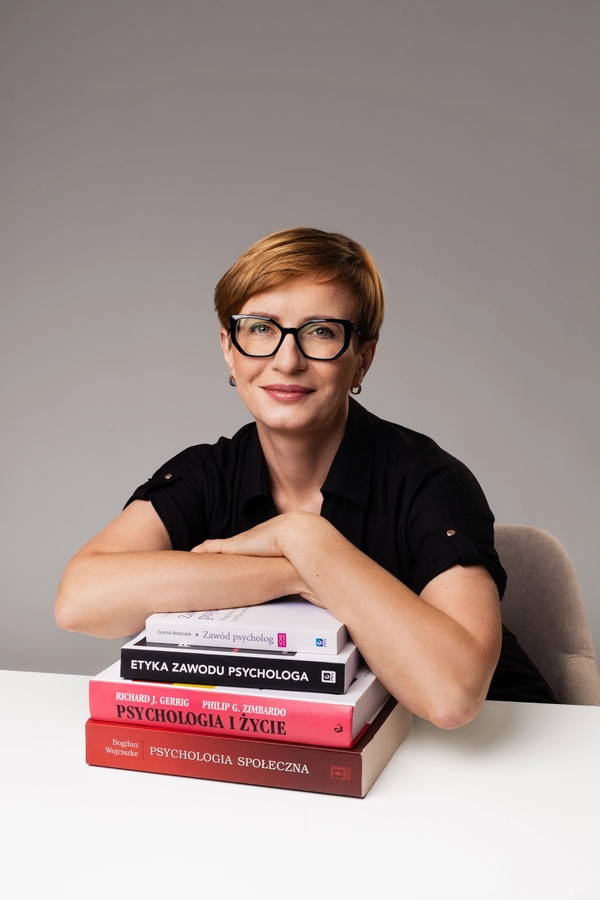Portret Lidii Rodowicz, która siedzi przy białym stole i opiera ręce o stos książek psychologicznych, w tym "Zawód psycholog", "Psychologia i życie" oraz "Psychologia społeczna".